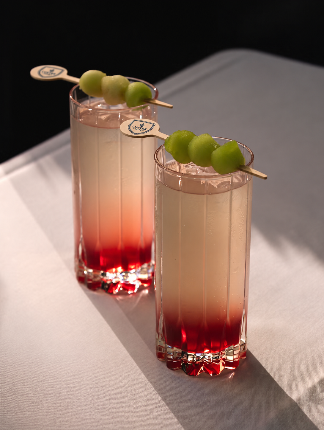 Two cocktails in cut-crystal glasses on a white tablecloth, with Luster Caffè picks